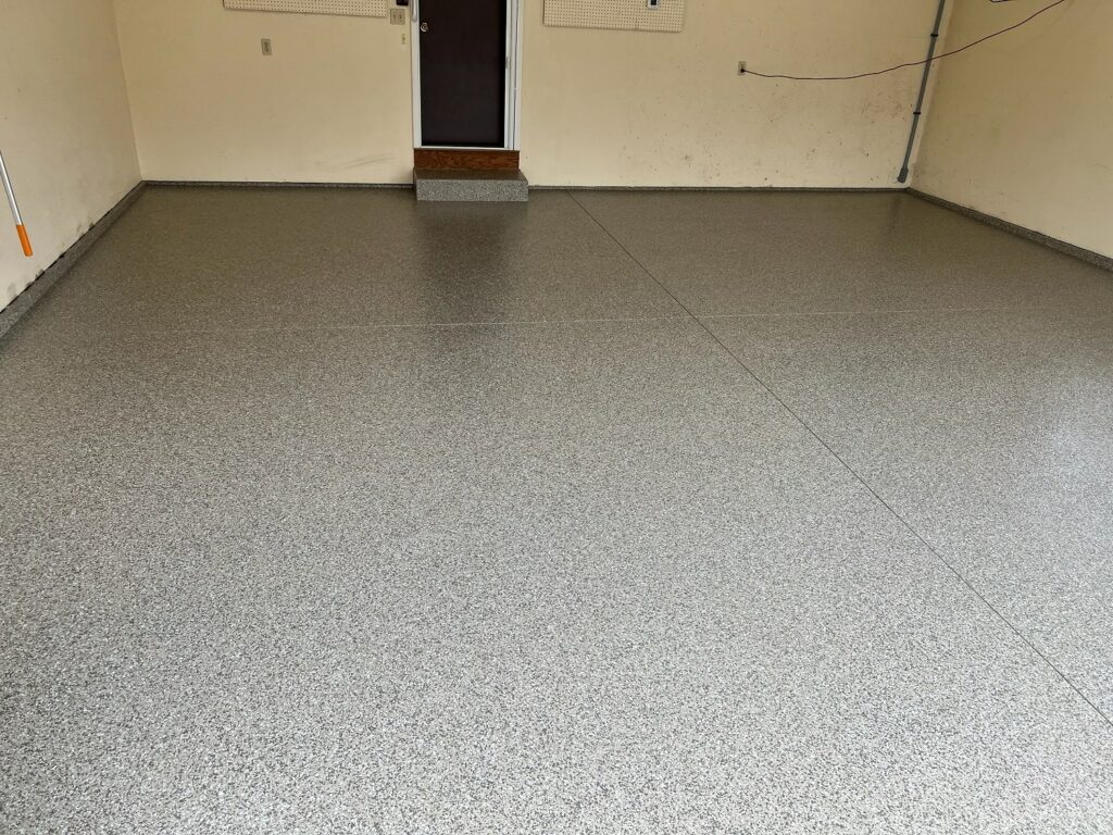 An empty room featuring a speckled grey floor with a dark mat before a closed brown door. Walls have a few wires and an electrical panel.