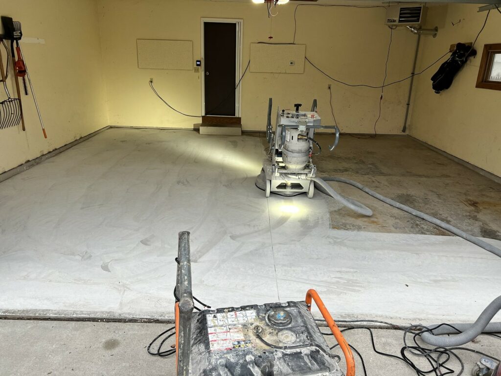 A concrete floor is partially covered with a white protective sheet. Concrete grinding equipment with hoses is visible in a garage-like setting.