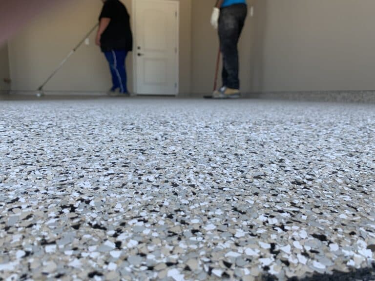 The image shows a speckled floor foreground with two people cleaning a room in the background, one sweeping while the other is standing.